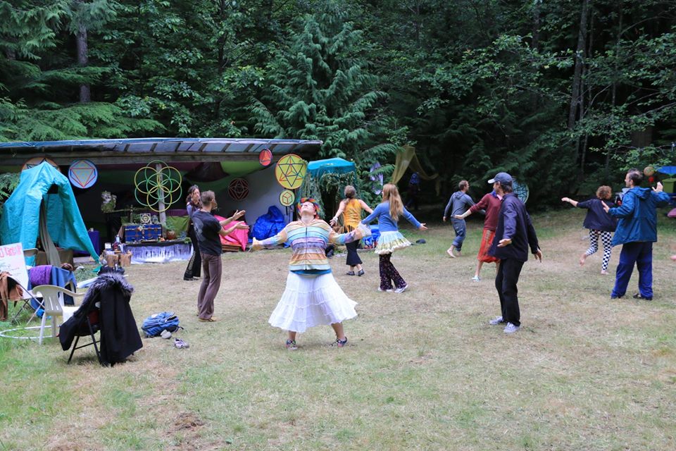 A group of people in a circle on a grassy field doing a movement exercise.  In the background is art, and altar and sound equipment.